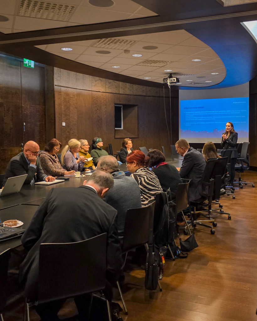 Joukko ihmisiä istumassa suuren pöydän ympärillä tummasävyisessä kokoushuoneessa.