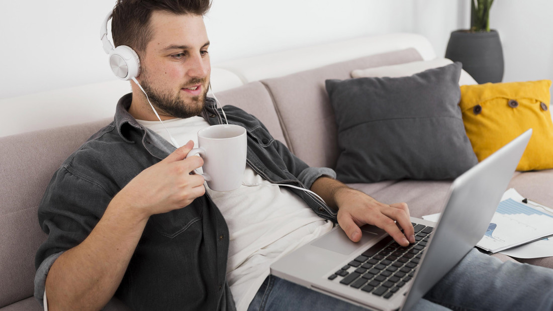 Mies istuu sohvalla luurit päässä, kahvikuppi kädessä ja tietokone sylissä.