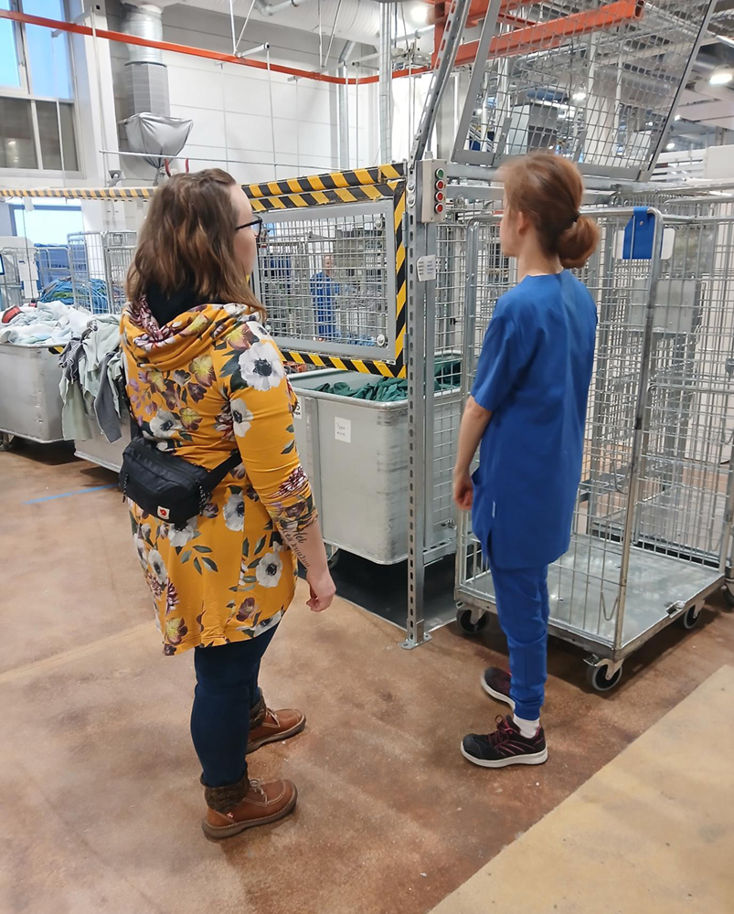 Kaksi naista suuressa pesulassa, jossa paljon pyykkihäkkejä. Toinen työasussa ja toinen arkivaatteissa.