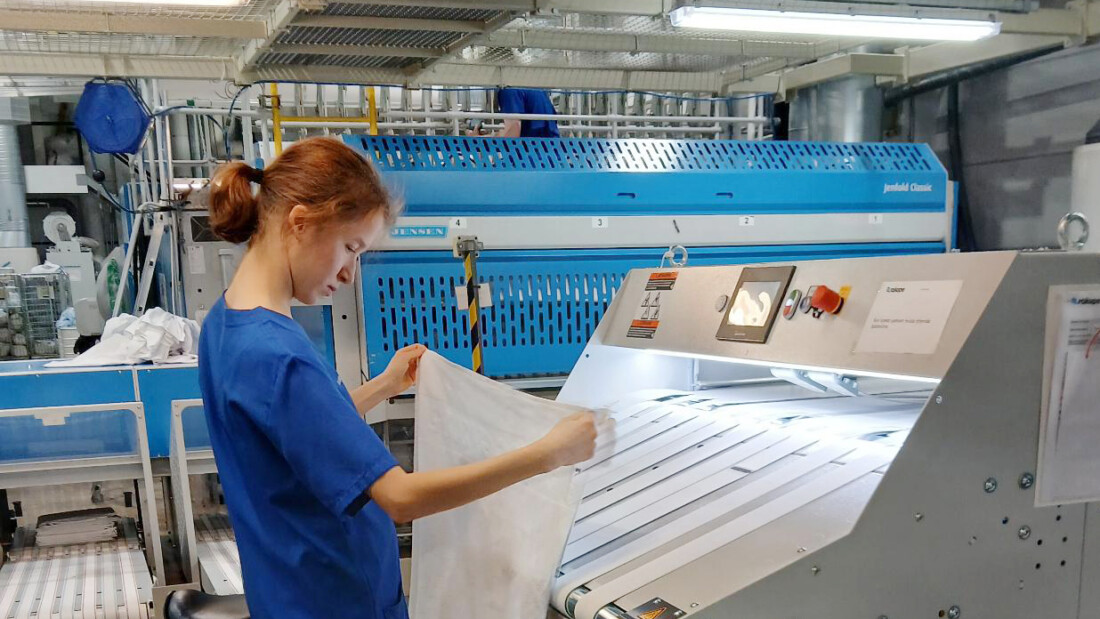 Henkilö työskentelee viikkauskoneen äärellä suuressa, hallimaisessa pesulassa.