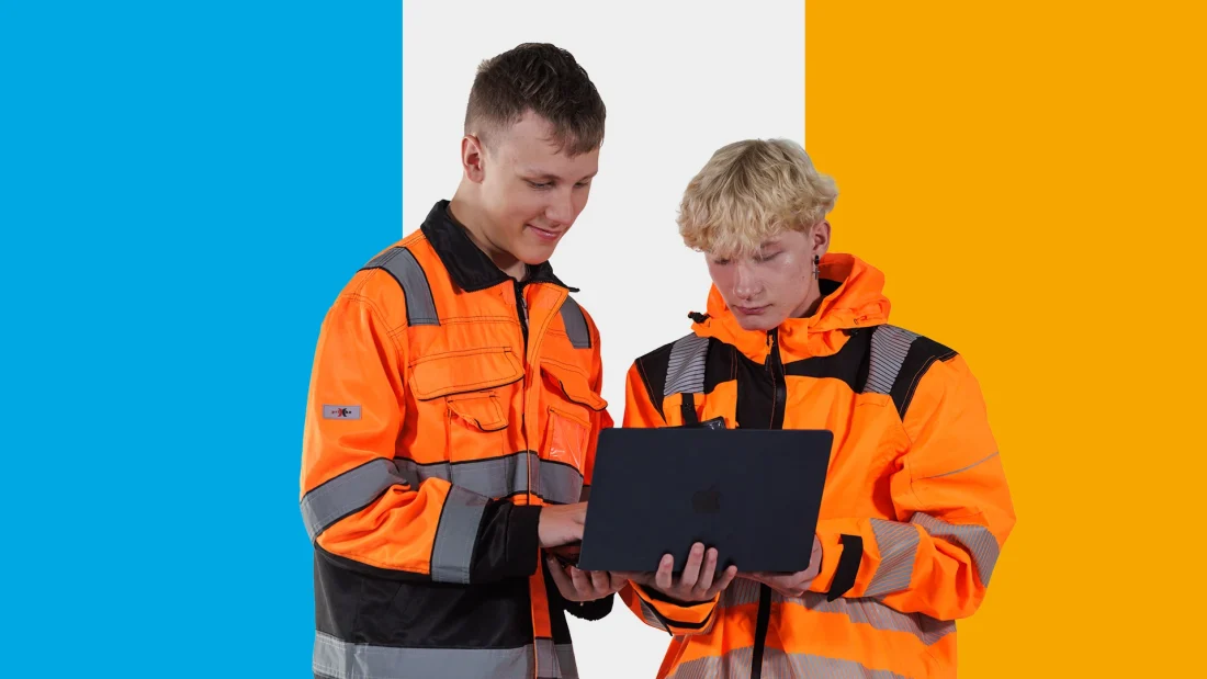 Kaksi oranssiin työasuun pukeutunutta nuorta tutkii tablettia yhdessä.