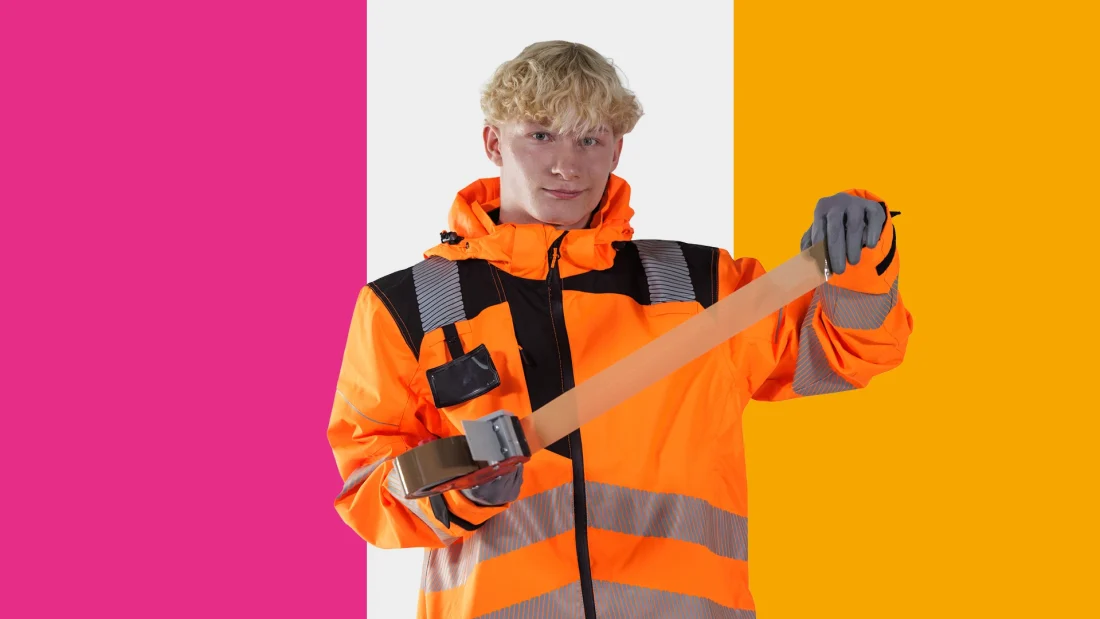 Oranssiin työtakkiin pukeutunut nuori vetää pakettiteippikoneesta teippiä esiin.