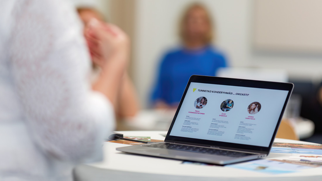 Tietokoneen näyttö ja sumeasti kaksi henkilöä, toinen etualalla ja toinen kauempana huoneessa.