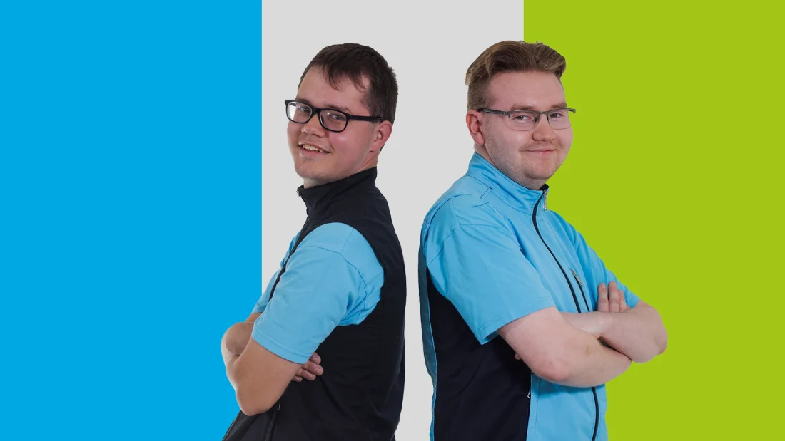 Kaksi puhtaanapitoalan työvaatteisiin pukeutunutta miestä seisoo selät vastakkain ja katsoo hymyillen kameraan.