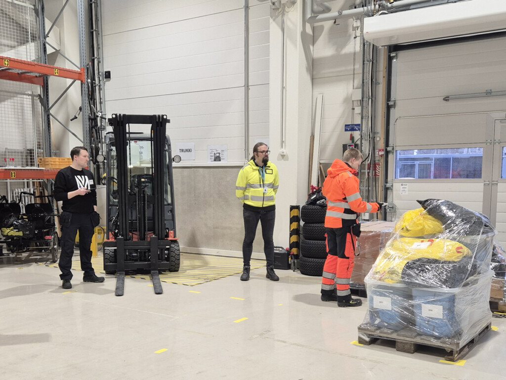 Kolme henkilöä logistiikkahallissa, yksi suorittaa tehtävää kuormalavan luona ja kaksi seuraa.
