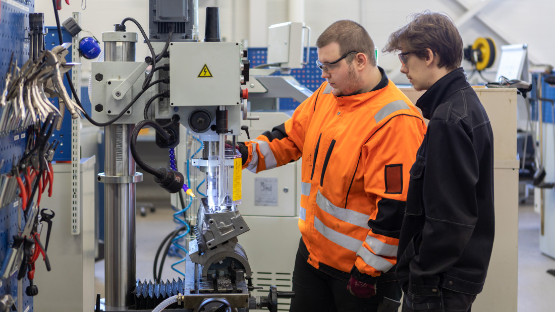 Kaksi koneasentaja-opiskelijaa työvaatteissa työskentelemässä koneen kanssa.