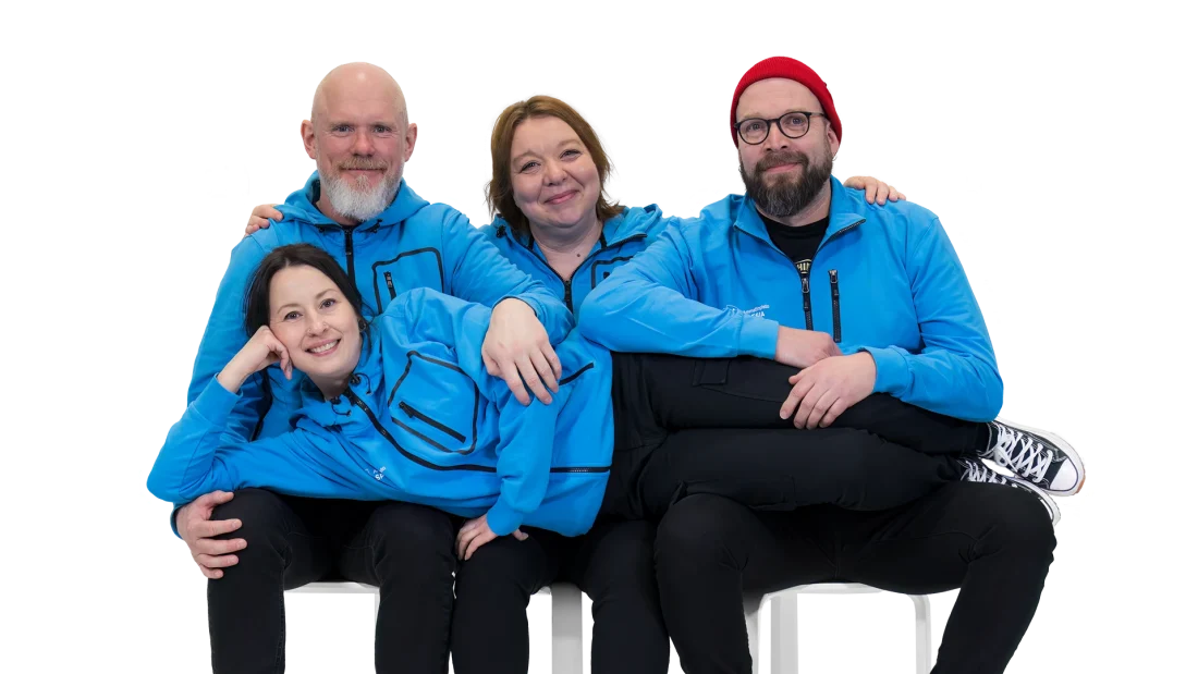 Kolme Spesian sinisiin takkeihin pukeutunutta aikuista istuu vierekkäin jakkaroilla ja neljäs henkilö makaa rennosti heidän polvillaan. Tunnelma on hauska.