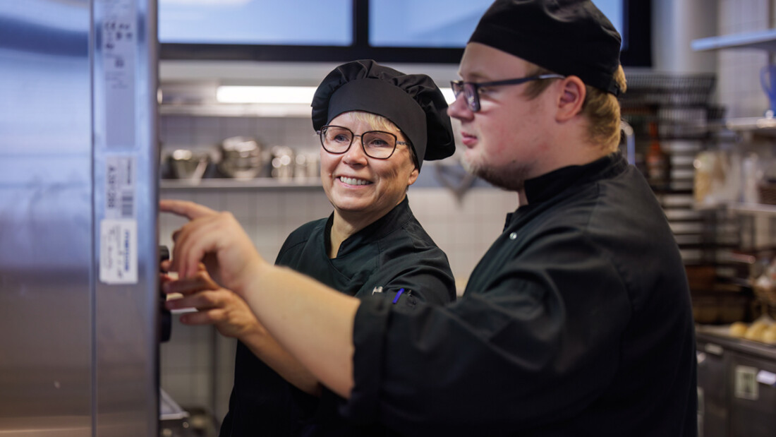 Kaksi tummiin kokkiasuihin pukeutunutta henkilöä suurtalouskeittiöllä, ohjaustilanne.