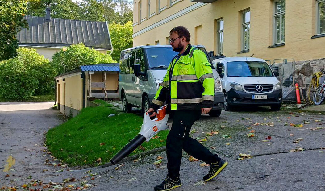 Spesian opiskelija Joonatan ulkona työvaatteet päällää puhaltamassa lehtiä lehtipuhaltimella rakennuksen edustalla.