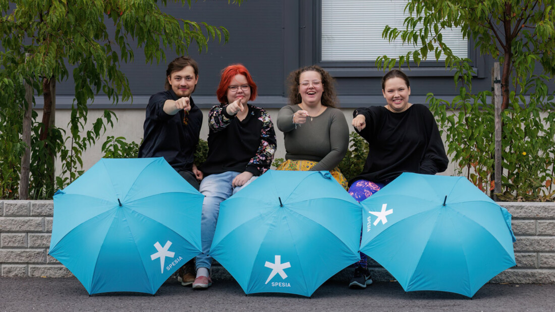 Neljä nuorta istuu tiiliaidalla, edessään kolme aukinaista Spesian sateevarjoa.