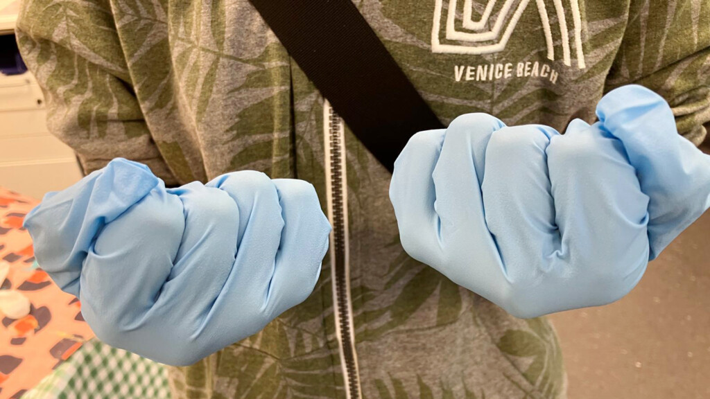 Lähikuvassa nyrkissä olevat kädet joissa siniset kumihanskat, käsien omistajalla päällään armeijanvihreä huppari.