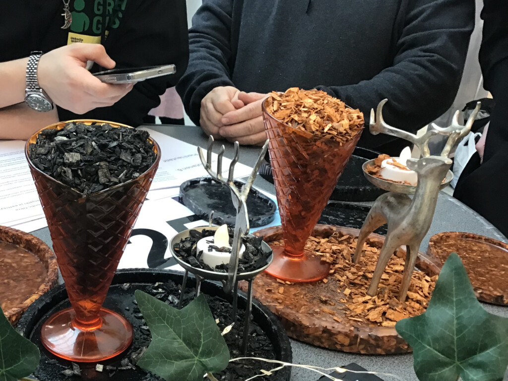 Lähikuva esittelypöydästä, joaa värjättyä haketta pikareissa sekä siitä tehtyjä puristetuotteita.