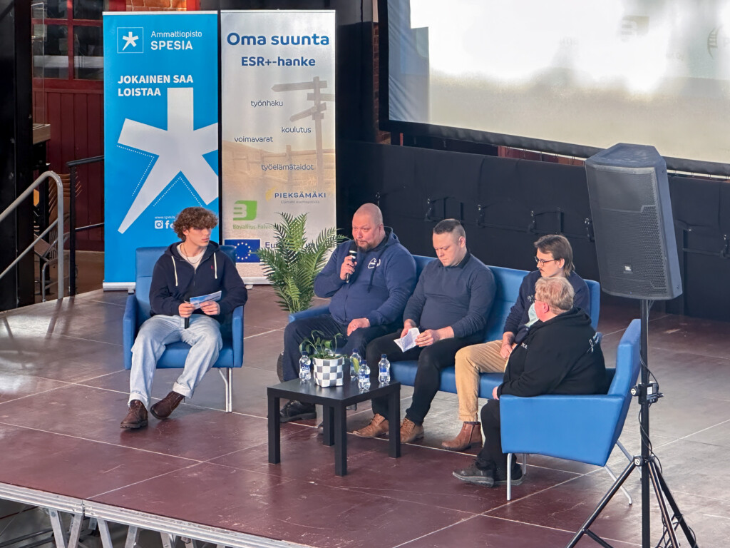 Neljä ihmistä nojatuoleissa tapahtumalavalla, taustalla Spesian roll-up.