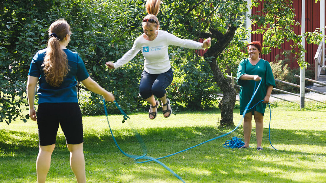 Kaksi henkilöä pyörittää ja yksi hyppii kesäisessä pihapiirissä.