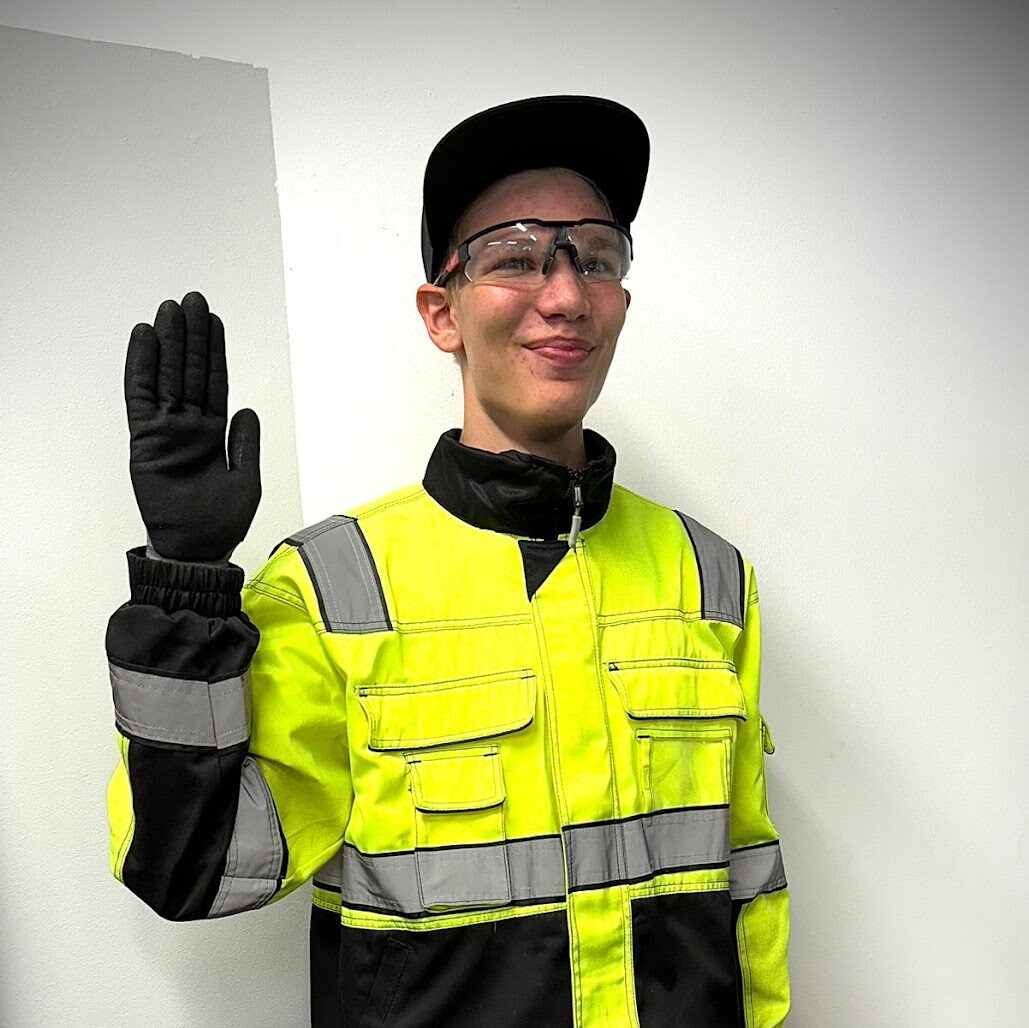 Henkilö työvaatteet päällään heiluttaa hymyilevästi.