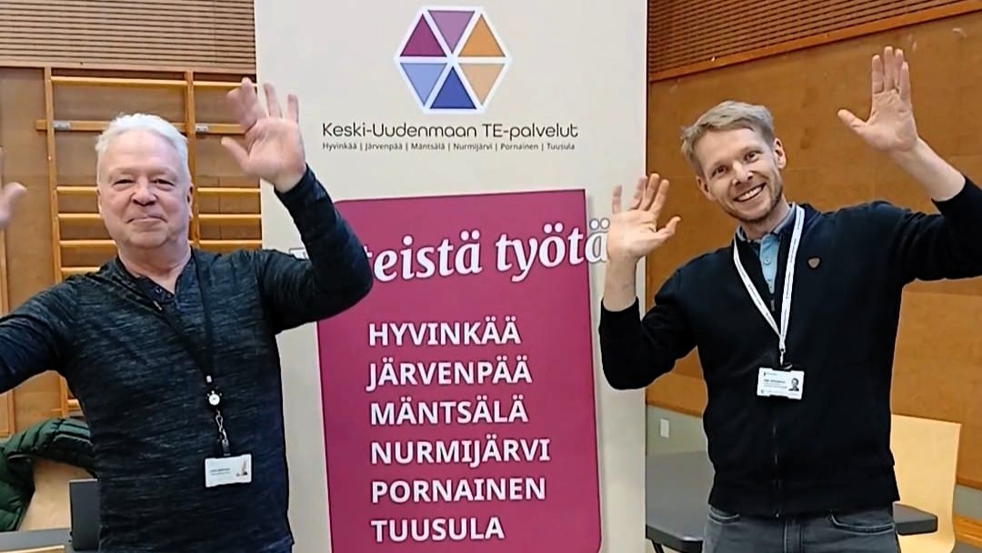 Kaksi henkilöä seisoo julisteen vieressä ja tervehtii käsillään iloisesti.