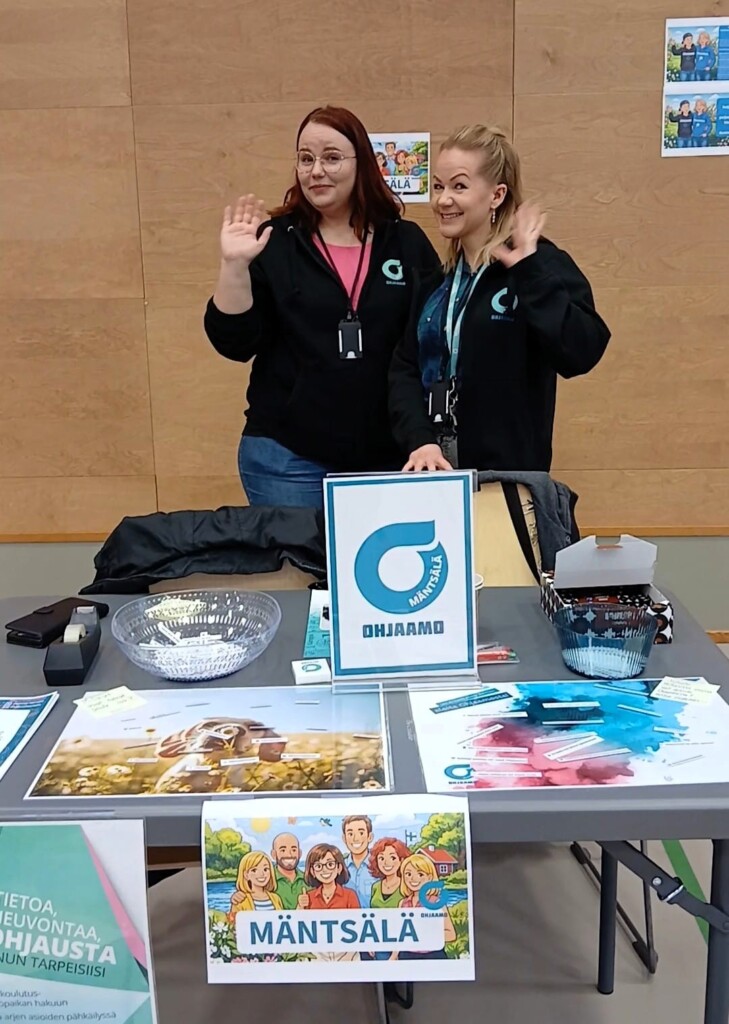 Kaksi henkilöä vilkuttaa esittelypöydän takaa. Pöydän päällä on esitteitä. 