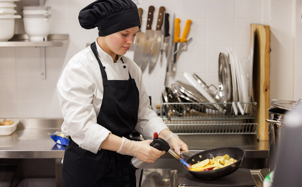 Nuori kokkiasussa liekittää paistinpannulla olevia omenasiivuja.