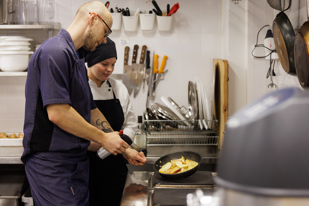 Opettaja opastaa kokkiasuiselle nuorelle liekittämistä.