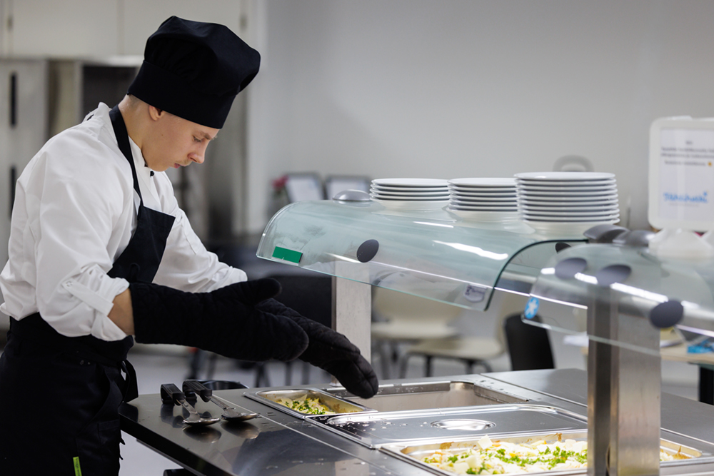 Kokkiasuinen nuori laittaa ruokaa esille linjastoon.