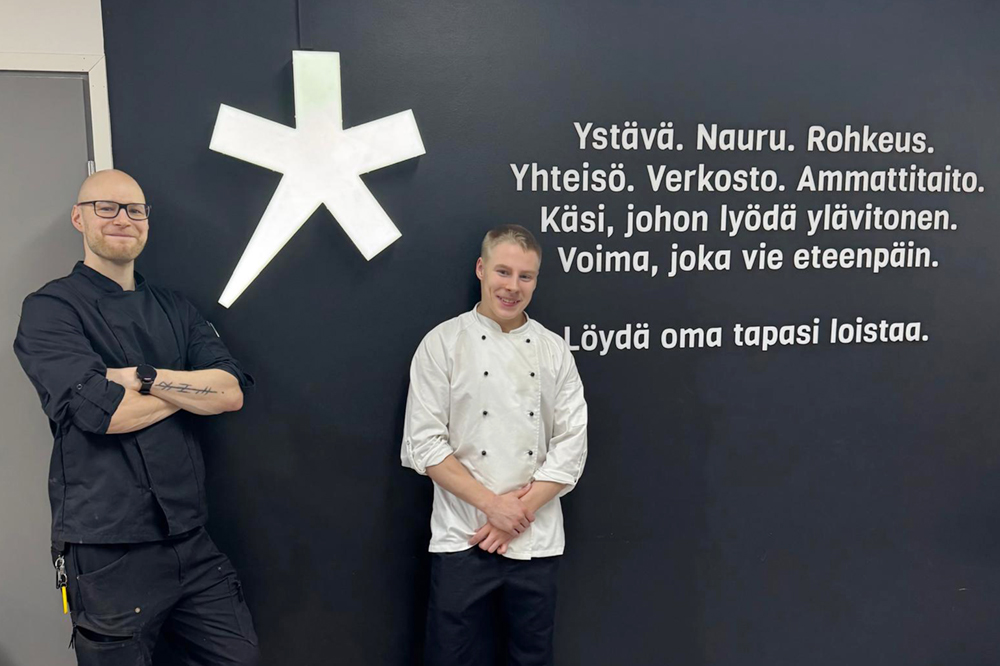 Kaksi keittiön työvaatteisiin pukeutunutta henkilöä seisoo Spesian voimalauseita sisältävän seinän edessä.