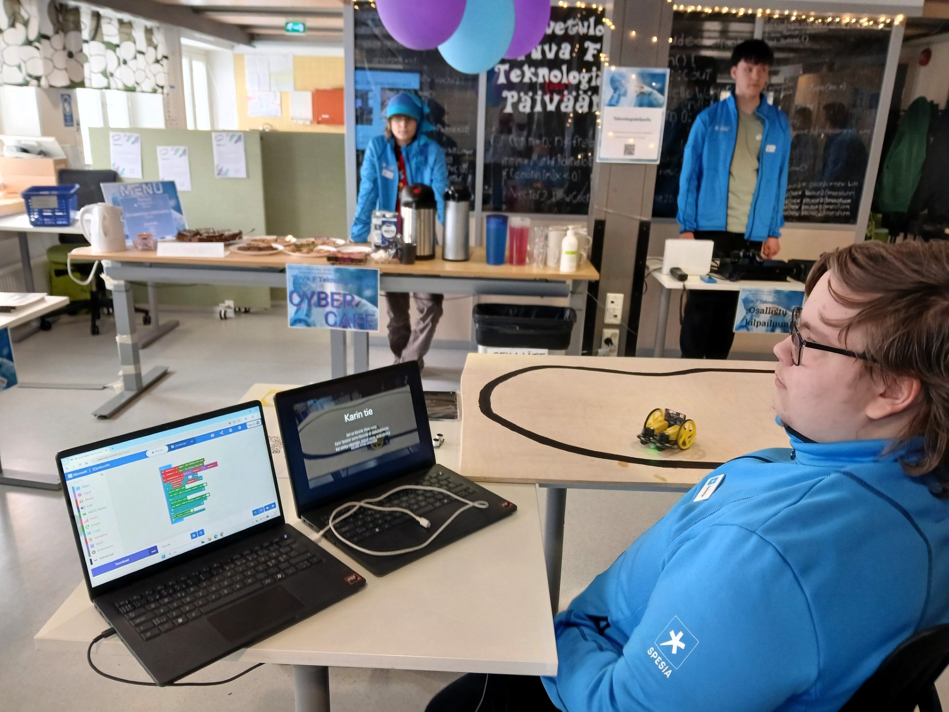 Opiskelijoita luokassa, jossa pöytiä. Etualalla kaksi kannettavaa tietokonetta, joiden on tekstiä näytöllä. 