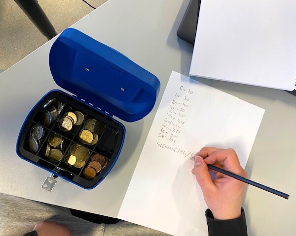 Pöydällä avoin kassalipas, jossa kolikoita ja henkilön käsi kirjoittamassa lukua paperille. 