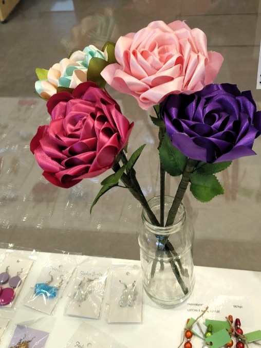 Silkkinauhasta tehtyjä ruusuja maljakossa pöydällä, jolla myös käsintehtyjä koruja. 