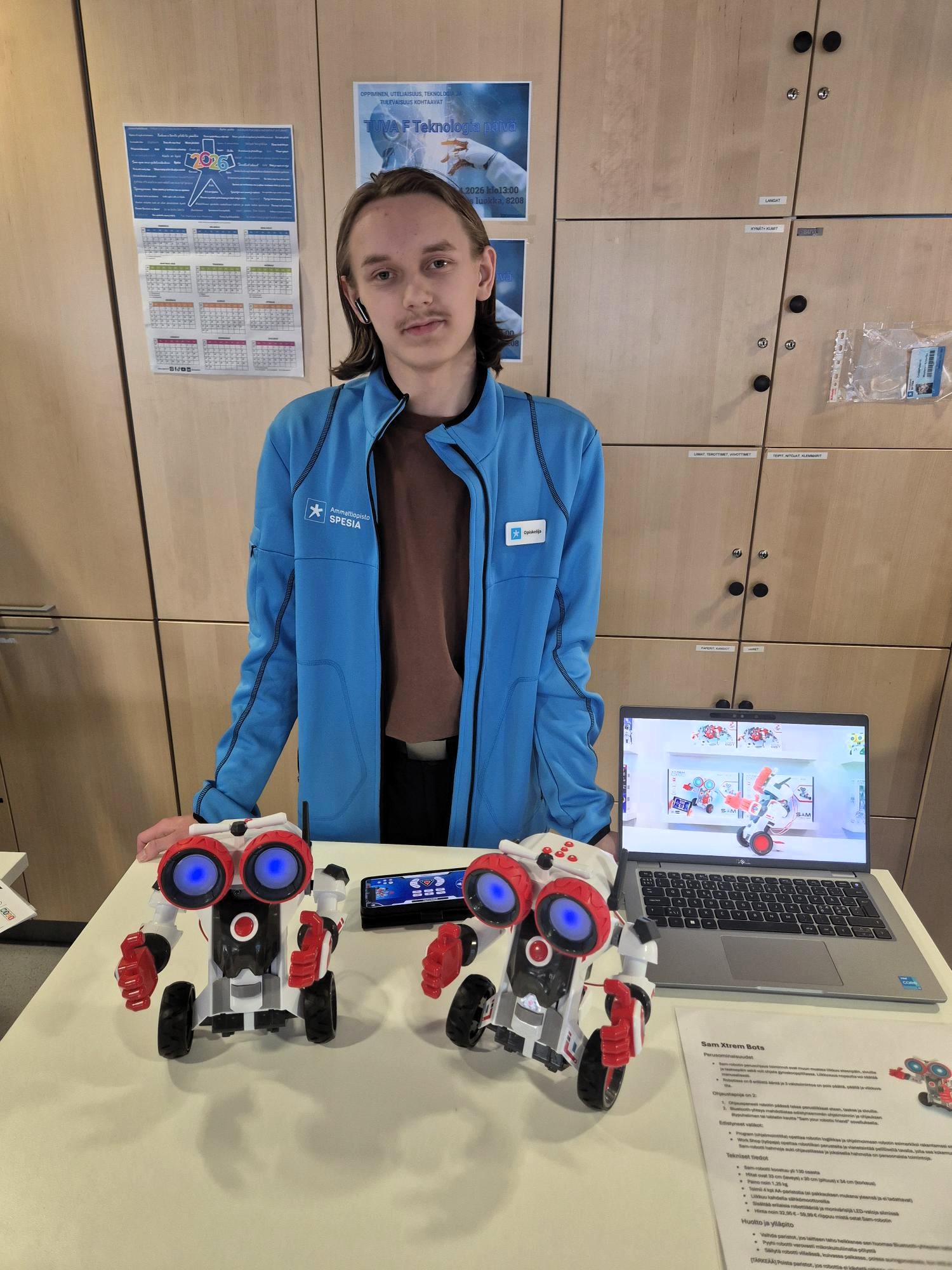 Opiskelija seisoo pöydän takana, jolle on asetettu kaksi robottia ja avoinna oleva tietokone. 