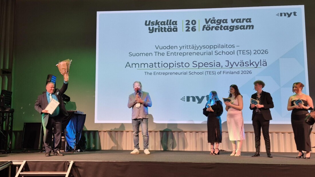 Kuusi ihmistä seisoo rivissä lavalla ja yhdessä on kädessään kunniakirja ja kukkakimppu.