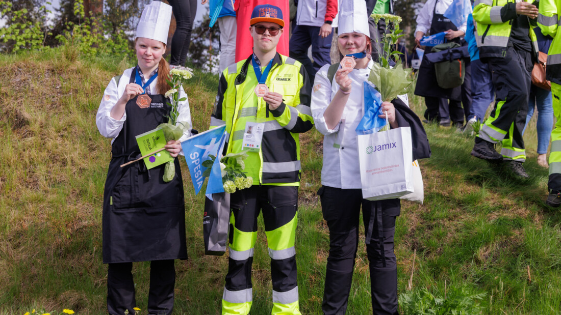 Kolme henkilöä seisoo nurmella pronssimitalit kaulassaan.