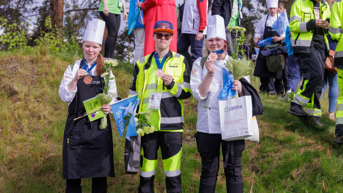 Kolme henkilöä seisoo nurmella pronssimitalit kaulassaan.