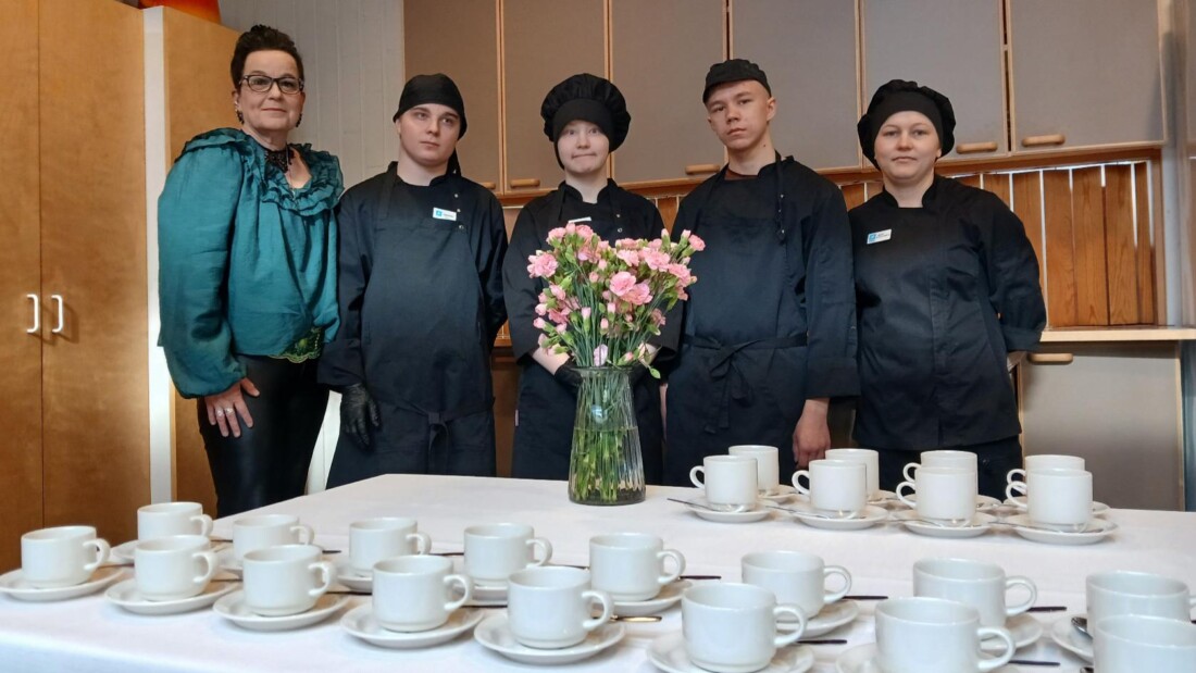 Kuusi henkilöä seisoo keittiötilassa pitkän pöydän takana. Pöytä on katettu valkoisella liinalla, kahvikupeilla ja aluslautasilla. Pöydän keskellä on maljakko, jossa on vaaleanpunaisia kukkia. Taustalla näkyy kaapistoja ja keittiövälineitä.