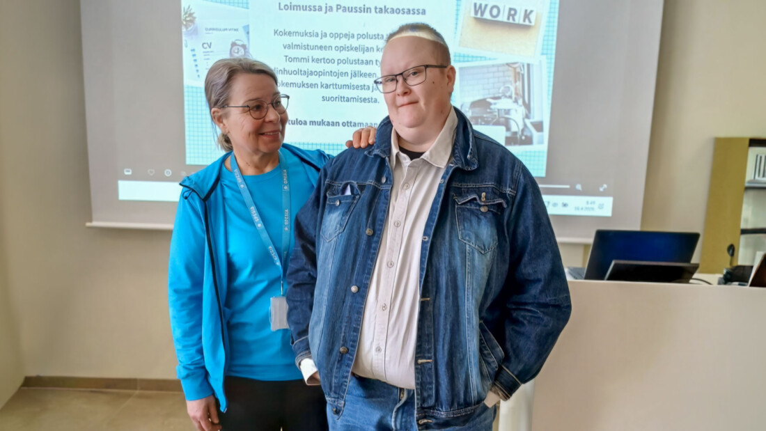 Kaksi henkilöä seisoo luokan edessä vierekkäin. Taustalla seinälle heijastettu esitys.