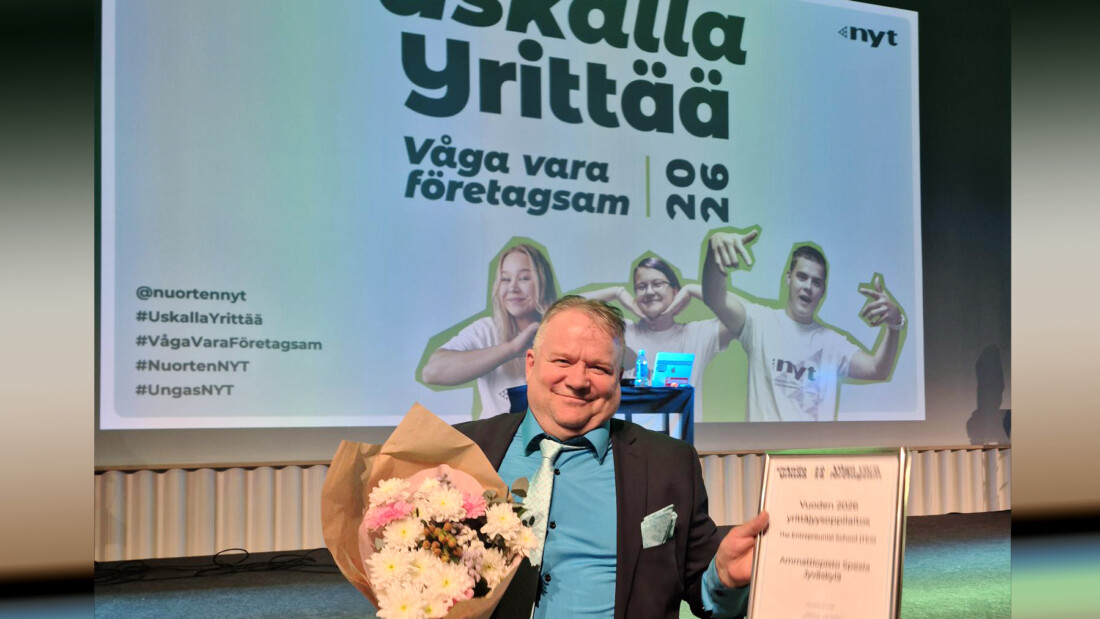 Opettaja Hannu Pahajoki kukkakimpun ja kehystetyn NYT-kunniakirjan kanssa, taustalle heijastettu Uskalla Yrittää -tapahtumakuva.