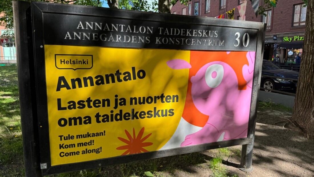 Annantalon taidekeskuksen ulkomainos Helsingin keskustassa. Kyltissä lukee "Annantalo – Lasten ja nuorten oma taidekeskus". Kyltti sijaitsee puiden varjostamalla alueella, ja taustalla näkyy katu sekä rakennuksia.