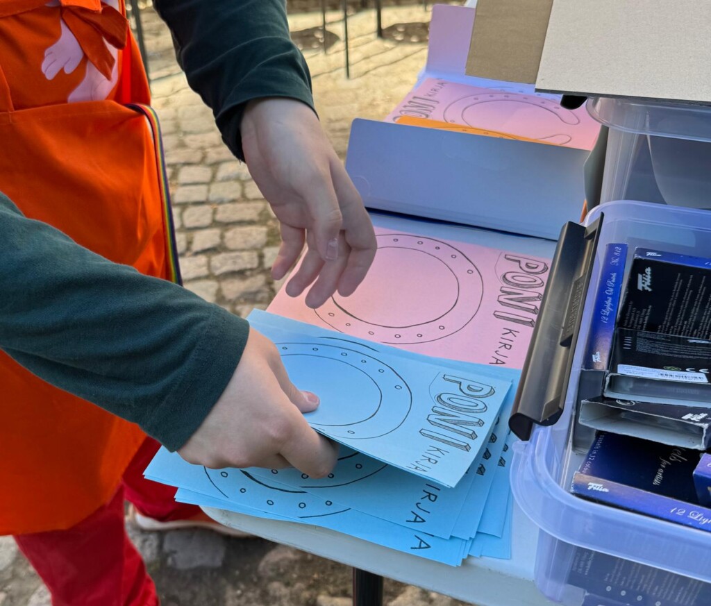 Henkilö lajittelee vaaleansinisiä ja vaaleanpunaisia tehtäväkortteja ulkona olevalla pöydällä. Kortteihin on painettu teksti ”PONI-kirja” sekä ympyränmuotoinen kuvio.