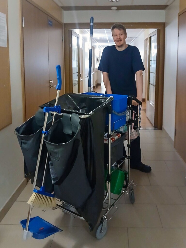 Opiskelija työskentelee siivousvaunun kanssa palvelukodin käytävällä. Vaunussa on siivousvälineitä ja jäteastioita.