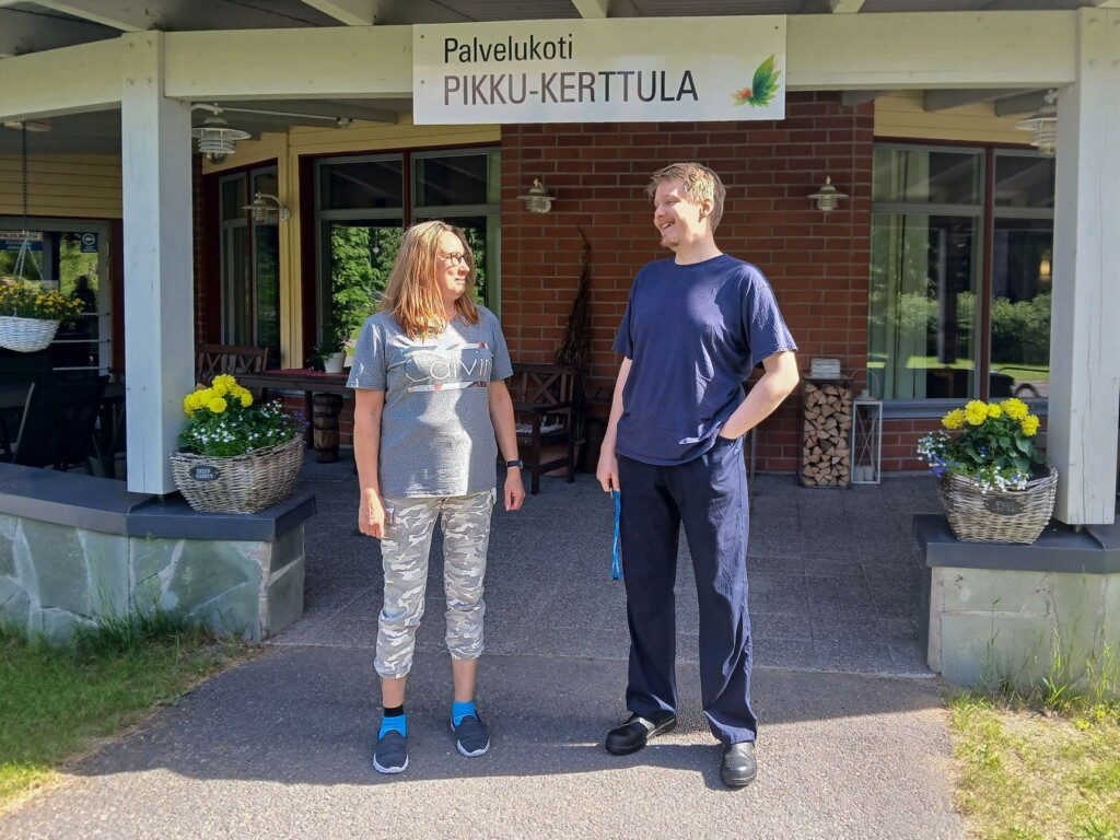 Kaksi henkilöä seisoo Palvelukoti Pikku-Kerttulan sisäänkäynnin edessä. Taustalla näkyvät rakennuksen katettu terassi, ikkunat ja kukkaistutukset.