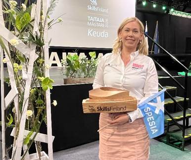 Spesian edustaja kilpailulavalla pitelemässä SkillsFinlandin järjestäjäkapulaa ja Spesian lippua Taitaja Mästare 2026 -tapahtumassa.
