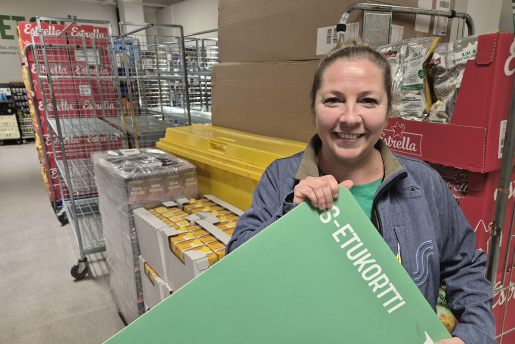 Myymäpäpäällikkö seisoo kaupan varastossa ja pitelee suurta S-etukorttia käsissään. Tutustu ja tienaa -esimerkki.