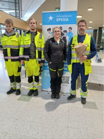 Neljä henkilöä seisoo Ammattiopisto Spesian roll-upin edessä kauppakeskuksen aulatilassa.