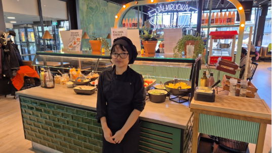 Henkilö seisoo ravintolan salaattibuffetin äärellä työvaatteissa ja päähineessä. Buffetpöydällä on tarjolla salaatteja, kasviksia ja lisukkeita.