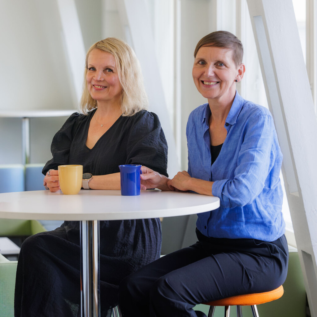 Tuula Niskanen ja Virpi Jussila istuvat pyöreän pöydän äärellä kahvikupit edessään.